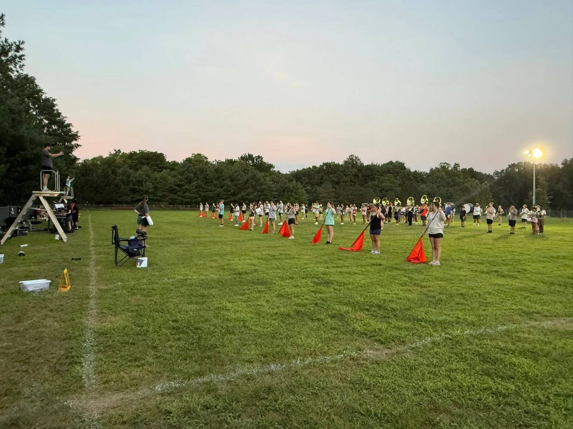 A marching band rehearses on a grassy field at dusk, with musicians and flag bearers arranged in rows and a conductor standing on a podium.