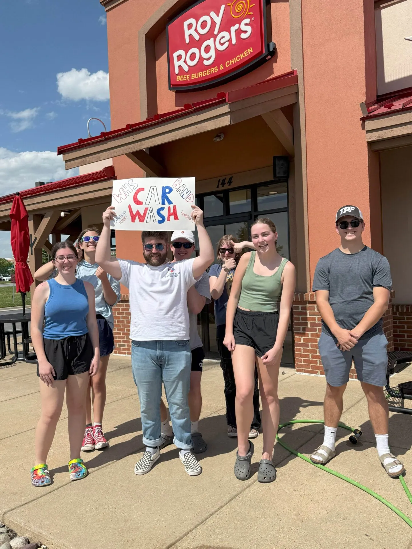 A group of six young people stand outside a Roy Rogers restaurant, one holding a sign that reads “Car Wash.” The weather is sunny and clear.