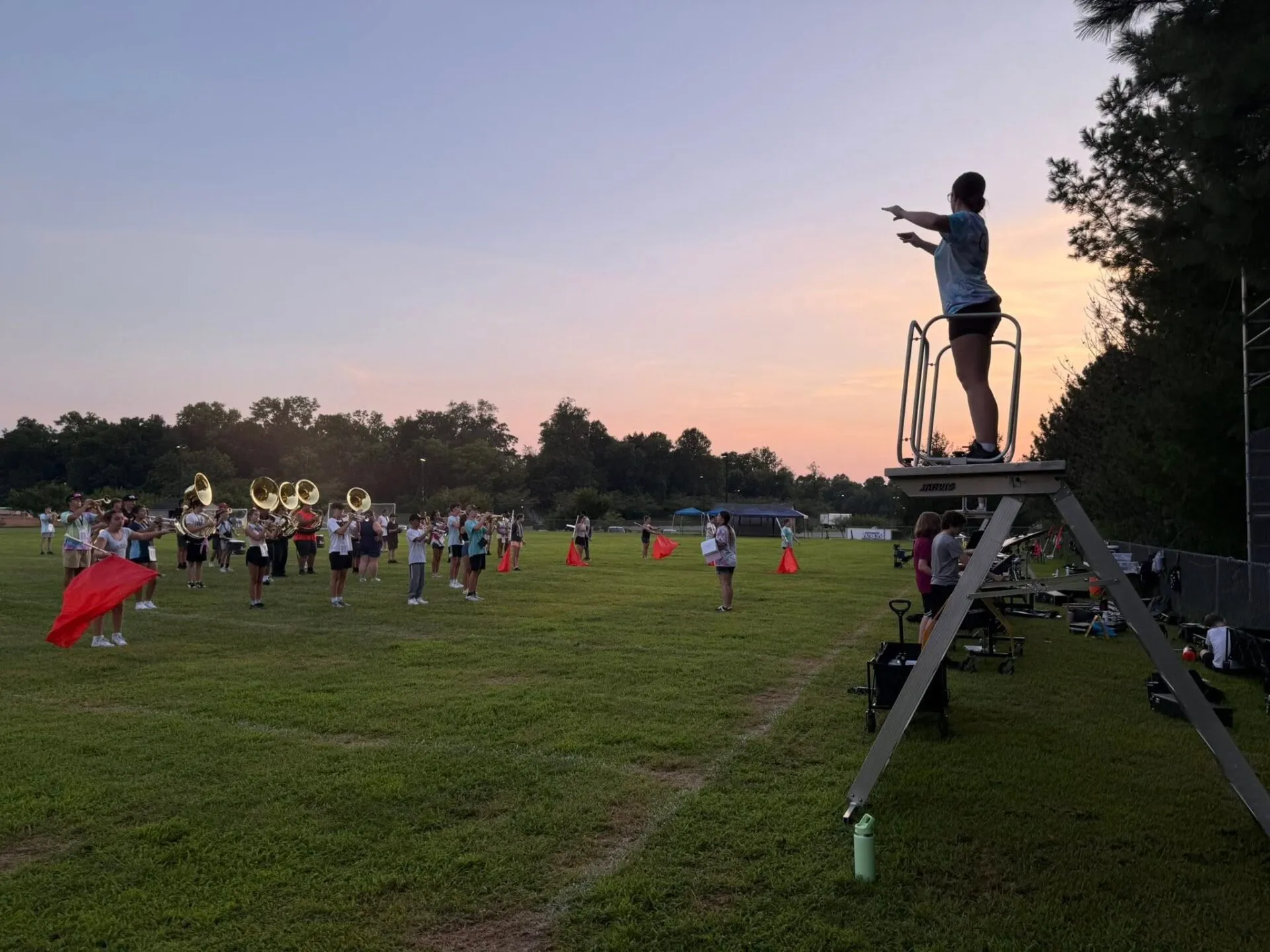 A marching band practices on a grassy field at dusk while a conductor stands on a tall metal podium, directing the group.